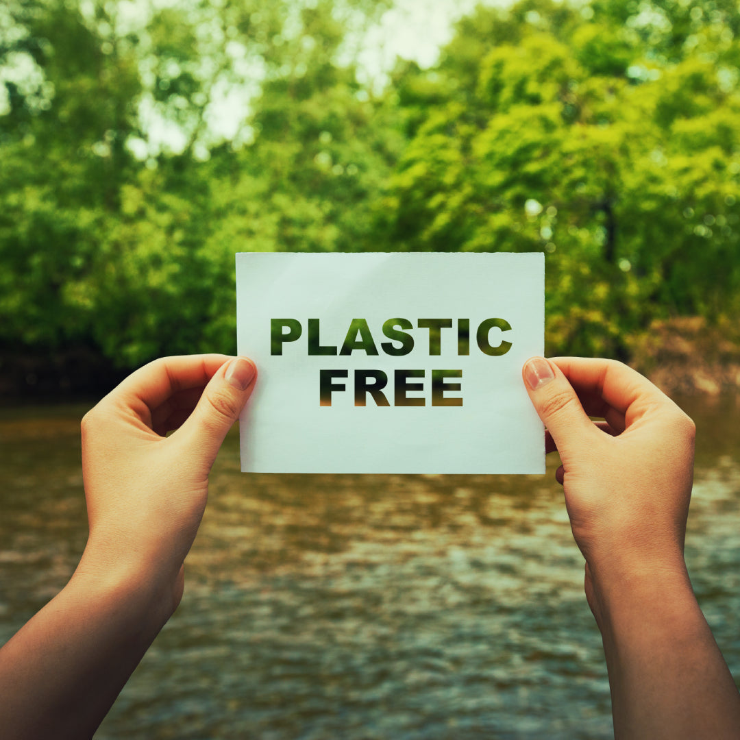 Hands hold a sign in front of trees lining a river. The sign reads 'Plastic Free'.